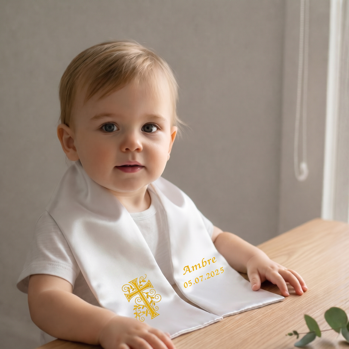 Illustration : bébé portant l'étole de baptême blanche brodée d'une croix avec feuillage dorée et du prénom « Ambre »