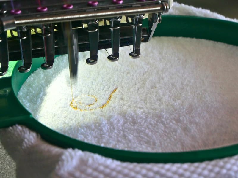 Illustration : brodeuse en action sur une serviette éponge blanche tendue dans un cadre de broderie vert