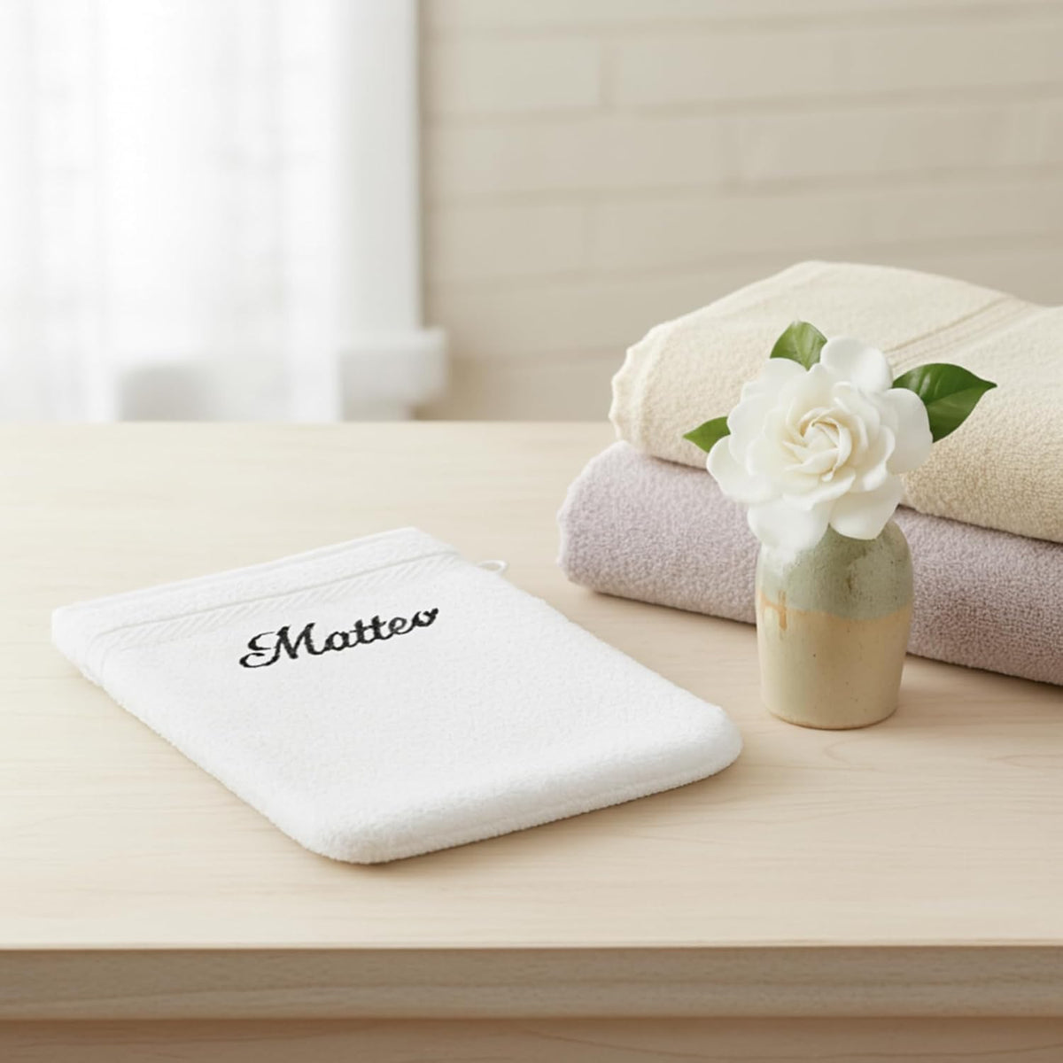 Gant de toilette blanc brodé « Matteo » posé sur un meuble, serviettes pliées et fleur blanche