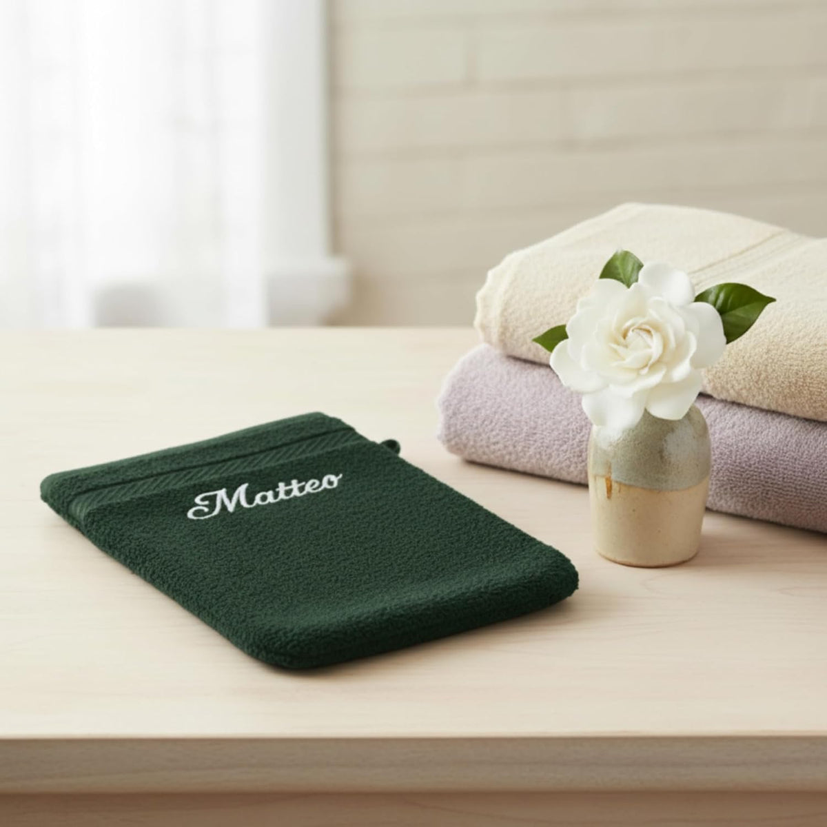 Gant de toilette vert foncé brodé « Matteo » posé sur un meuble, serviettes pliées et fleur blanche
