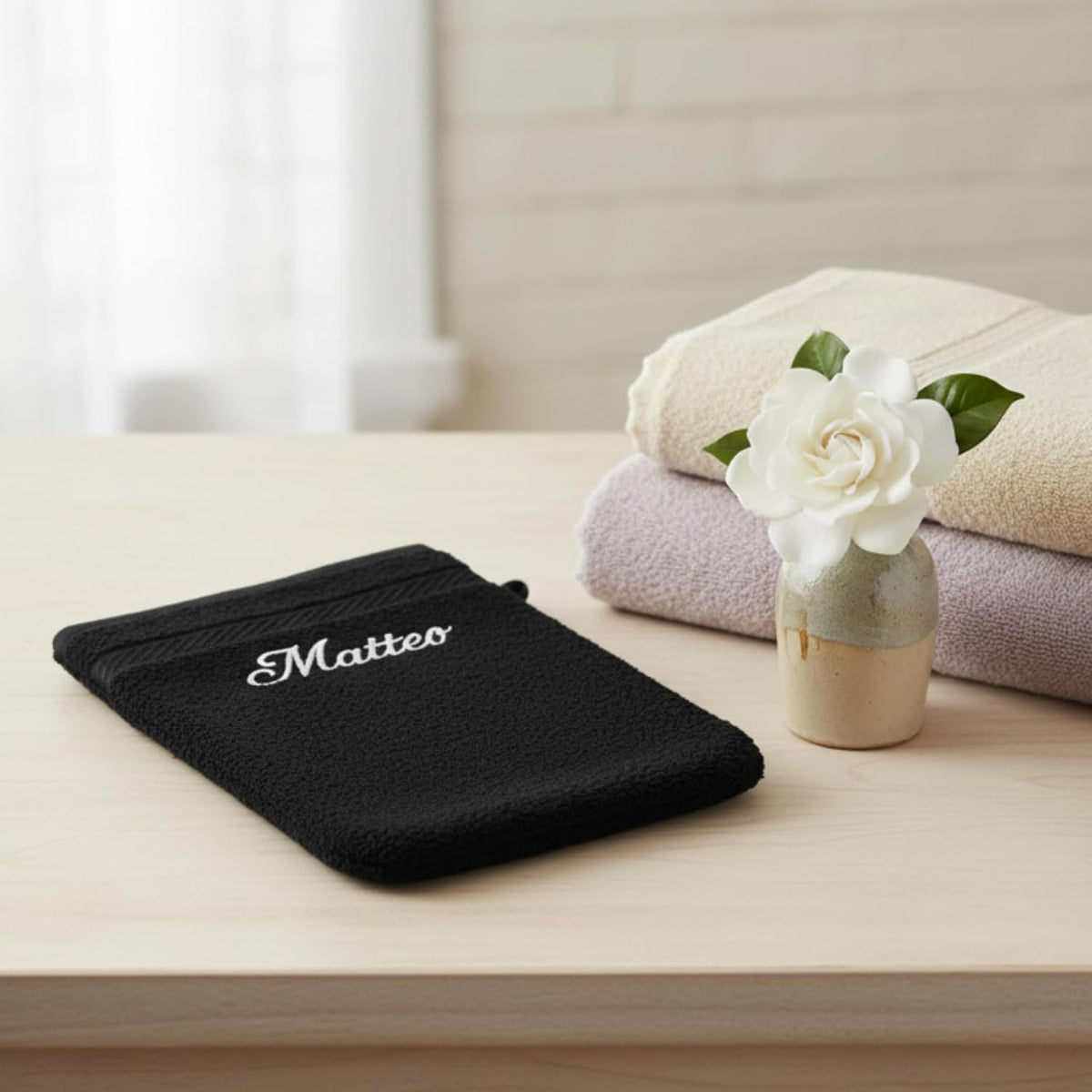 Gant de toilette noir brodé « Matteo » posé sur un meuble, serviettes pliées et fleur blanche