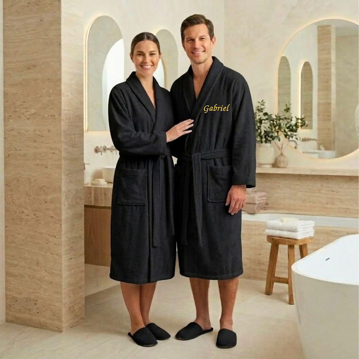 Couple en peignoirs noirs dans une salle de bain, celui de l'homme brodé « Gabriel » en fil doré