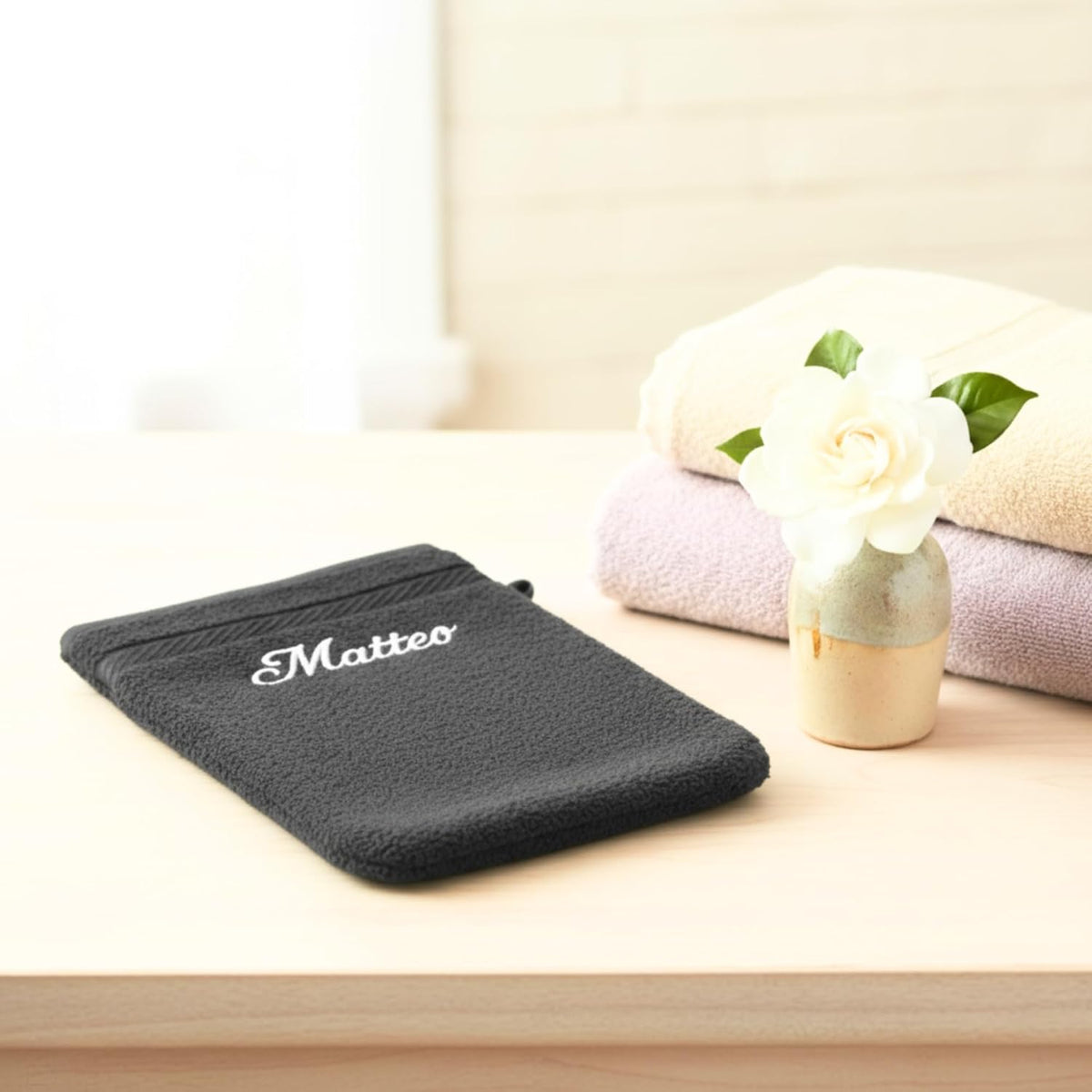 Gant de toilette anthracite brodé « Matteo » posé sur un meuble, serviettes pliées et fleur blanche