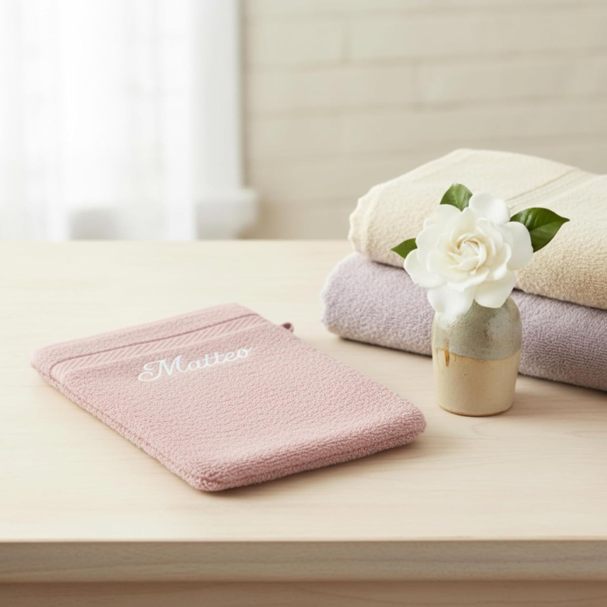 Gant de toilette rose brodé « Matteo » posé sur un meuble, serviettes pliées et fleur blanche