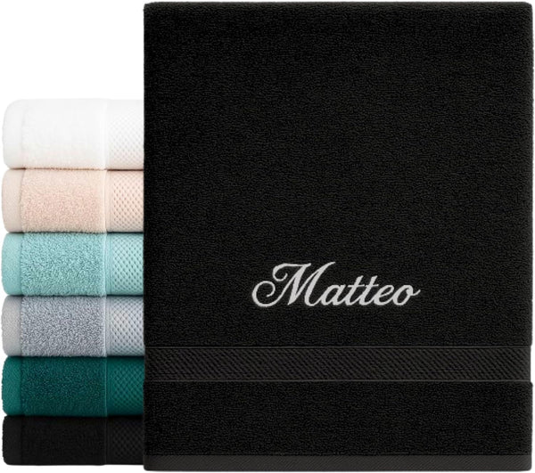 Serviette noire brodée du prénom « Matteo » en fil blanc, devant une pile de serviettes de couleurs