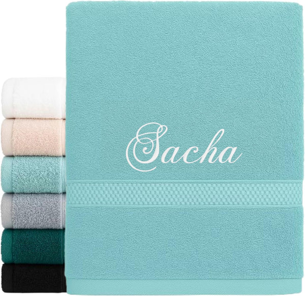 Serviette bleu turquoise brodée du prénom « Sacha » en fil blanc, devant une pile de serviettes
