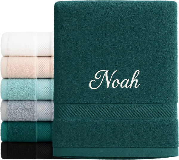 Serviette verte brodée du prénom « Noah » en fil blanc, devant une pile de serviettes de couleurs